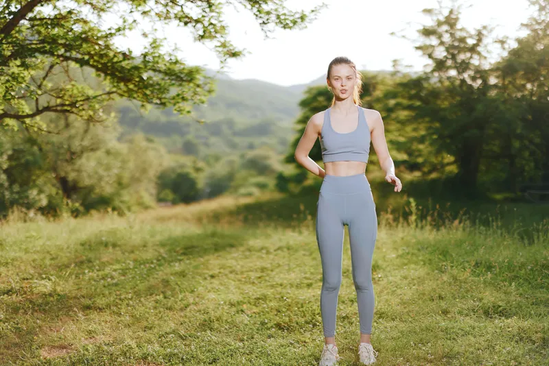 Young athletic woman in gray sportswear stands confidently in a serene green landscape, promoting fitness and healthy lifestyle choices in nature