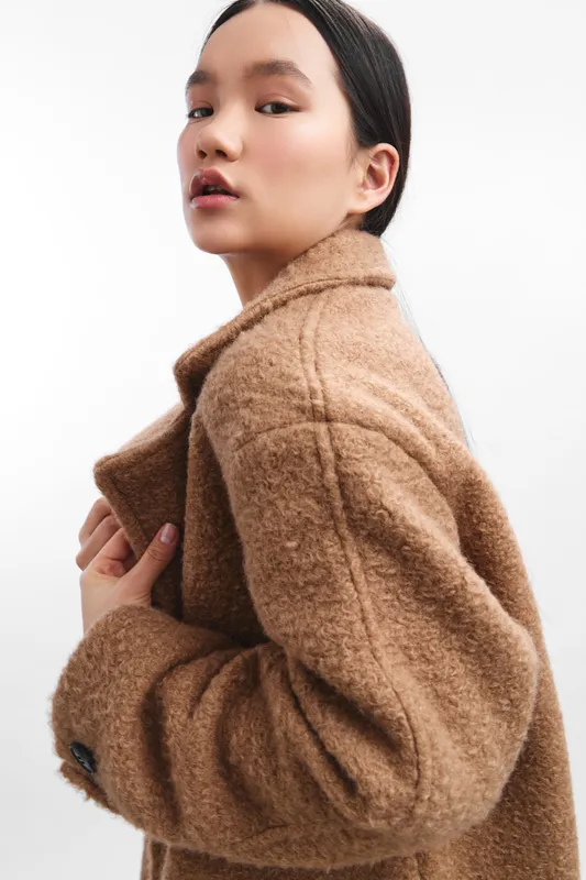 asian woman wearing a warm beige coat stands in a studio with a clean neutral background, conveying elegance, confidence, and contemporary style.