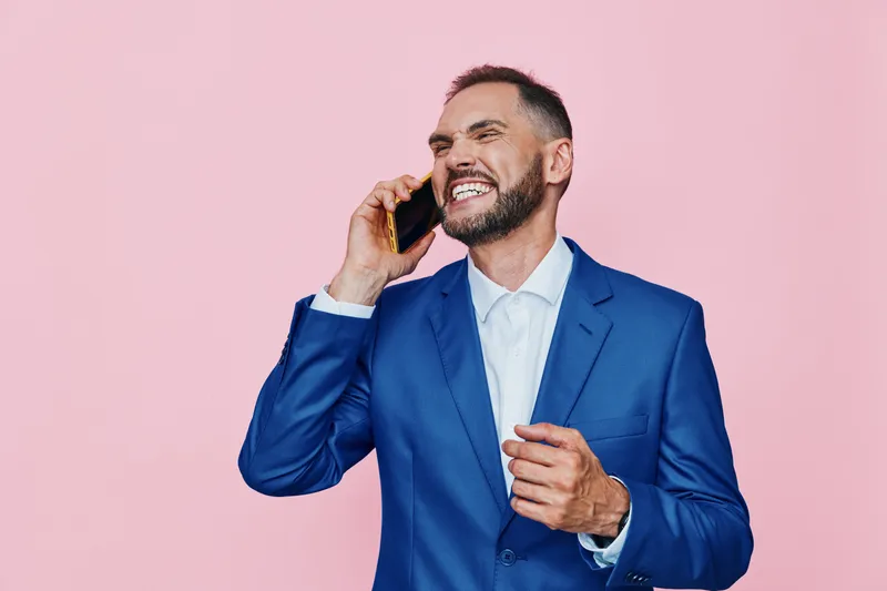 A professional man in a blue suit on a pink background speaks on the phone with a confident smile, showcasing business charisma, focus, and optimistic leadership.