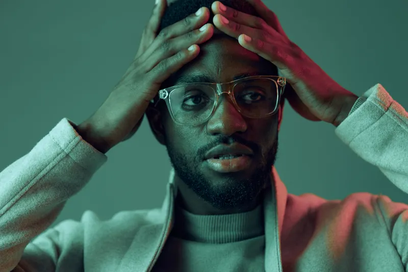 A contemplative Black man wearing glasses and a light jacket, displaying a thoughtful expression against a stylish green and pink background, suggesting introspection and emotional awareness