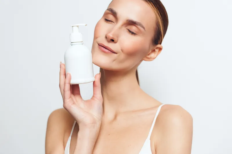 Woman in tank top holds pump bottle lotion to face, skincare routine with moisturizer, beauty and spa concept, calm expression, isolated on white background, gentle dermatology care today
