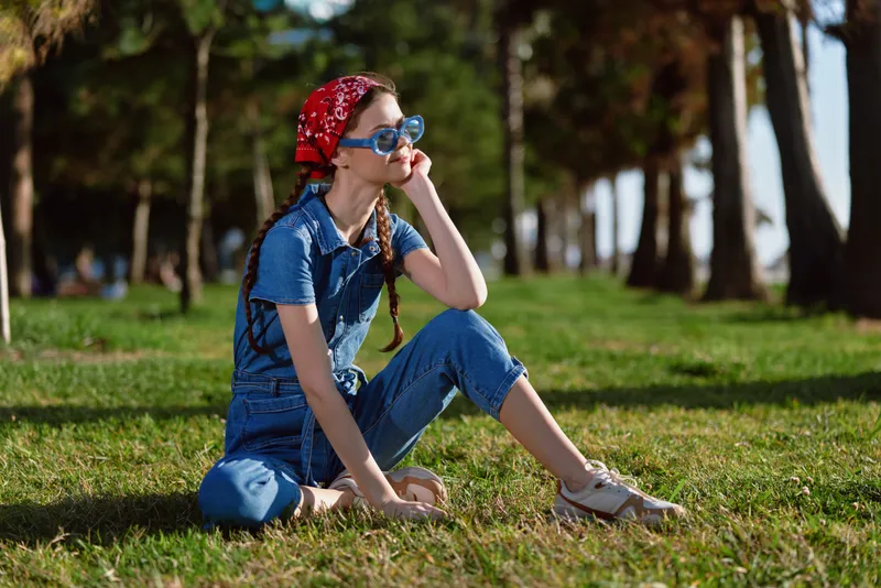 young woman sitting on grass wearing blue denim overalls and red bandana enjoying sunny day outdoors with sunglasses and thoughtful expression