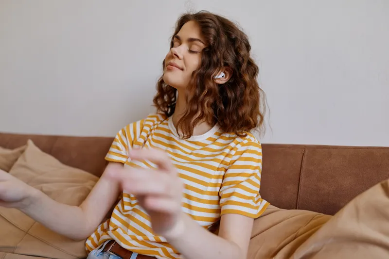 Bright woman indoors, relaxed on a couch, striped yellow shirt, sunlight streaming in, casual lifestyle moment, cozy home setting, natural colors, peaceful mood, with gentle breeze and cozy