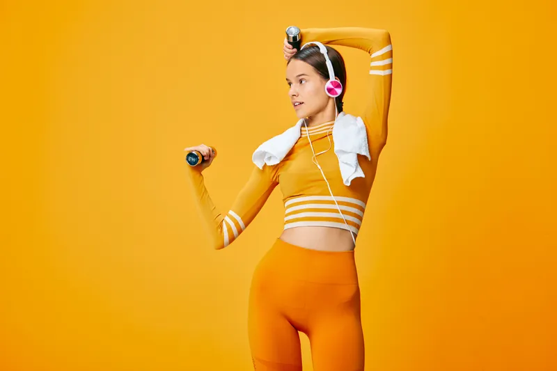 Woman in bright orange sportswear lifts a dumbbell, headphones on, towel around neck, in a bold orange studio. Dynamic fitness scene focused on strength, training, and athletic lifestyle.