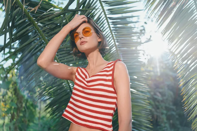 A stylish woman wears a red and white striped crop top and sunglasses as she poses outdoors amid lush palm fronds, creating a vibrant summer lifestyle scene.