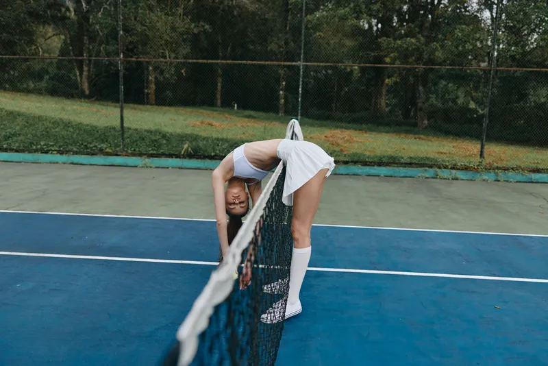 Young athletic woman in white sportswear stretching over a tennis net on a sunny day, showcasing determination and focus in a vibrant outdoor setting