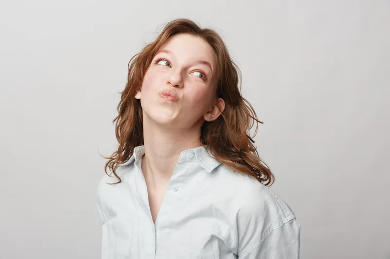 Young woman with red hair wearing white shirt looks up with playful expression on light background. Concept of fun, curiosity, and casual lifestyle portrait in studio lighting.