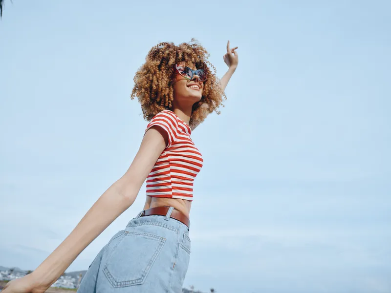 Joyful young woman with curly hair wears a red striped crop top and denim jeans, posing with arms outstretched under a clear blue sky for a carefree summer vibe.