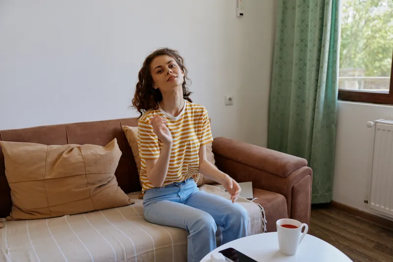Woman with curly hair sits on a couch at home, wearing a striped yellow shirt and jeans. Close, inviting scene suggesting casual lifestyle, comfort, and everyday relaxation in bright room.