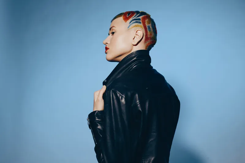 Woman with colorful shaved hair wearing black leather jacket, standing confidently against blue background in modern edgy style, expressive and bold appearance.