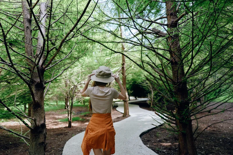 Woman walking on a curved path through a lush forest wearing a sun hat and orange skirt, back view of a traveler enjoying nature trail with trees, canopy and peaceful greenery.