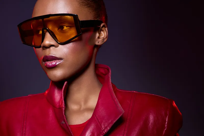 Portrait of a confident black woman wearing oversized orange sunglasses and a red leather jacket. Studio lighting highlights her smooth dark skin and stylish modern look.