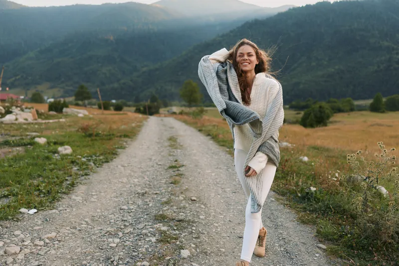 Young woman with long hair walks along a rural gravel road, enjoying the outdoors in a cozy shawl, smiling under a cloudy sky.