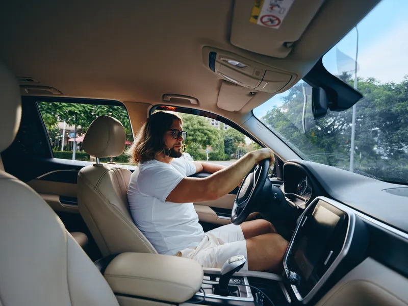 A man with long hair drives a sleek modern car, wearing a white tee. The bright interior, steering wheel, and calm roadside view convey a relaxed travel vibe.