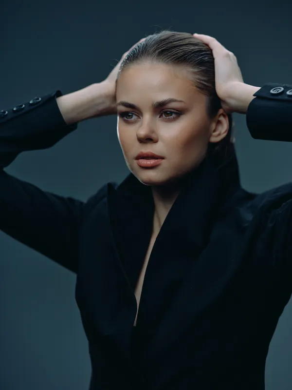 Portrait of a stylish woman with slicked-back hair wearing a black coat, showing a thoughtful expression against a dark background.