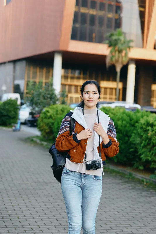 Woman with backpack and camera stands outdoors in casual urban fashion, ready for street photography and travel, jeans and jacket, sunny day near modern buildings