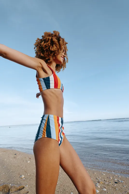 Young woman with curly hair wearing colorful striped bikini standing on the beach with arms outstretched. Clear sky and calm sea in the background create a peaceful summer mood.