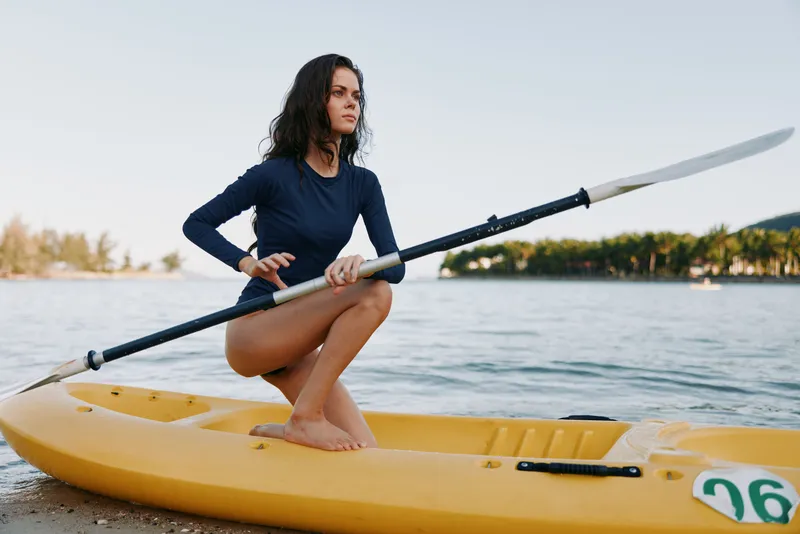 A young woman kneels on a bright yellow kayak near the shoreline, gripping a paddle and gazing toward the horizon, capturing balance, focus, and outdoor adventure on calm water.
