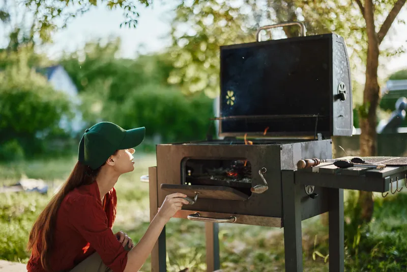 Young woman preparing barbecue outdoors, wearing casual attire and a cap, enjoying the sunshine in a vibrant green garden setting
