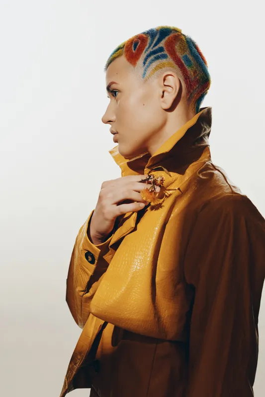 Woman with colorful shaved hair wearing a mustard-yellow textured jacket, posing in profile against a clean white background with a thoughtful expression.
