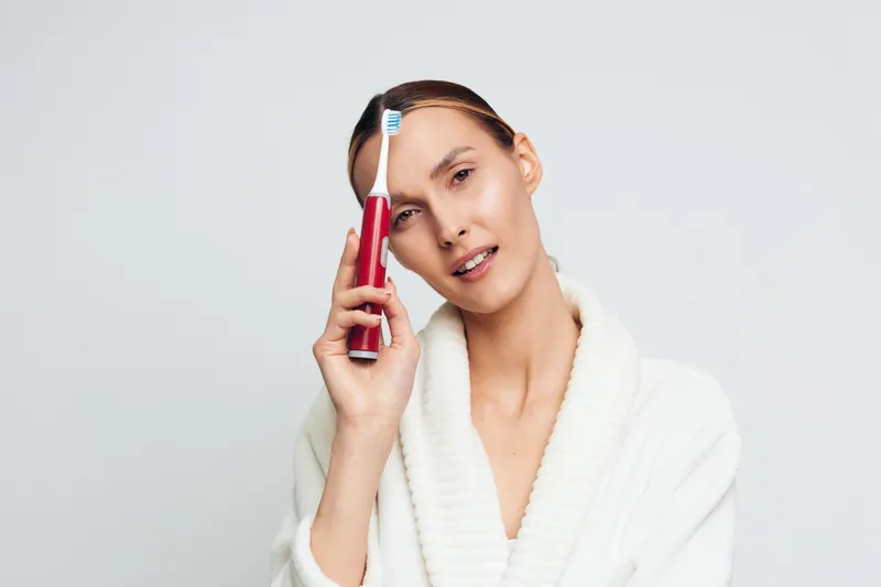 Young woman in white bathrobe holds red electric toothbrush near face with gentle smile isolated on white background for dental hygiene concept and personal care routine.