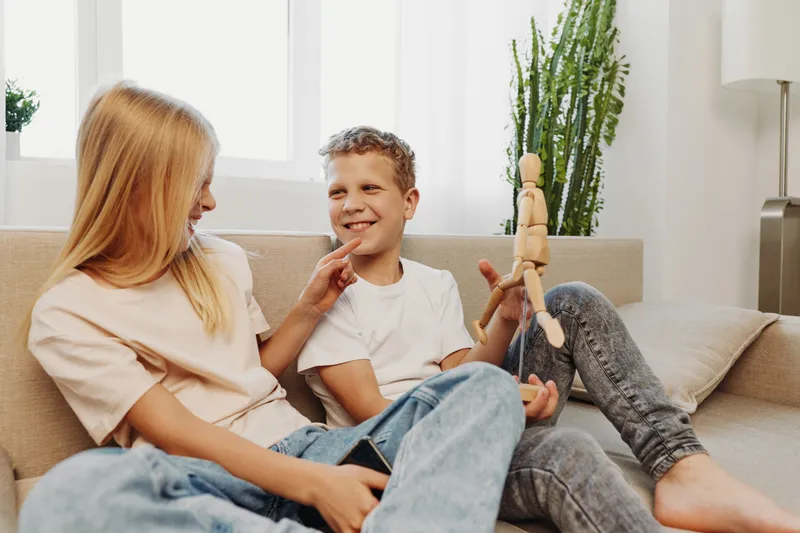 Happy siblings sitting on sofa at home, smiling and playing with wooden mannequin toy. Cozy indoor family moment with natural light and casual clothes.