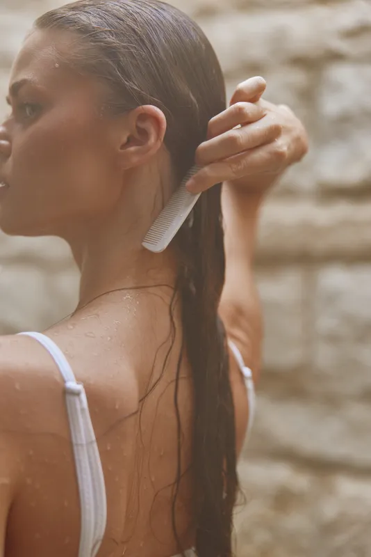 woman combing wet hair with white comb under natural light, showing delicate skin and water droplets, with blurred neutral background and soft tones