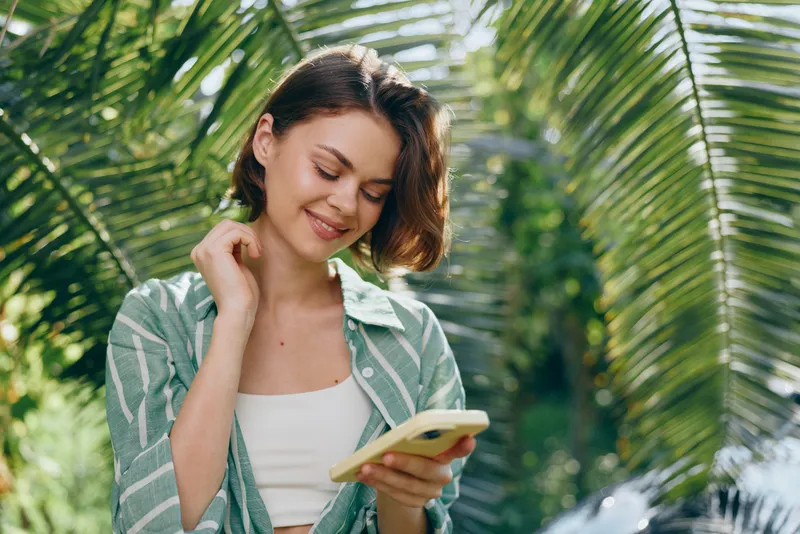 Woman reading a book or tablet outdoors among lush palm leaves, soft sunlight, relaxed pose, and a gentle smile in casual green attire.