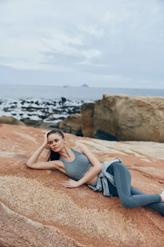 Young woman in activewear lounging on rocky beach, looking relaxed and confident, with ocean in background, emphasizing fitness and serenity