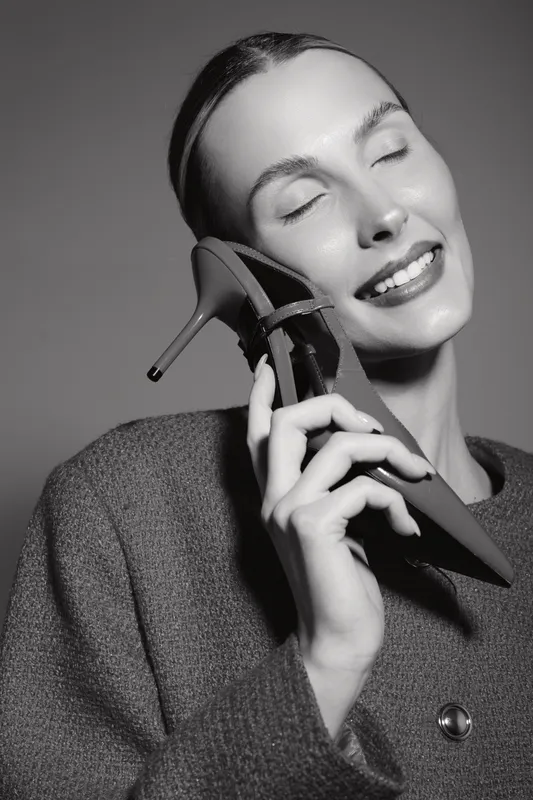 Happy caucasian woman with light skin smiling with closed eyes holding high heel shoe near face, wearing sweater against plain background. Concept of fashion joy and style.