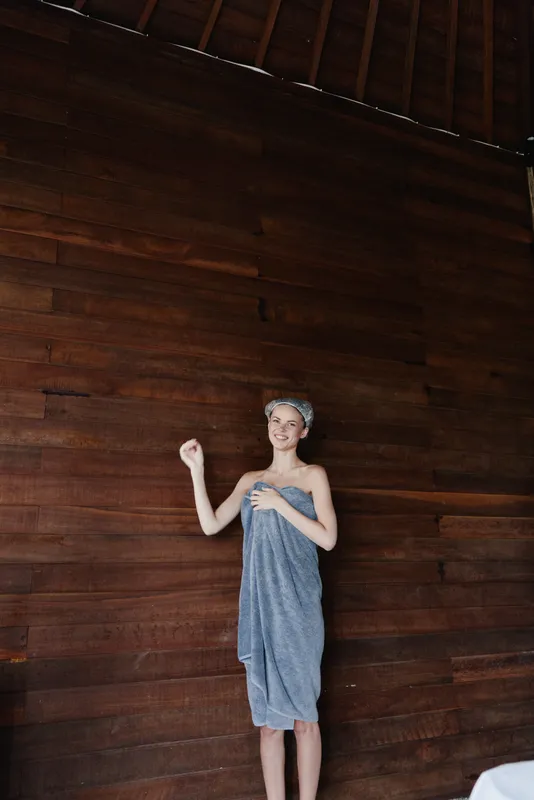 smiling woman wrapped in blue towel with headband stands against dark wooden wall showing joyful and relaxed mood in spa or wellness environment
