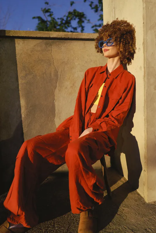 Cheerful woman outdoors in a bold red outfit, seated against a sunlit wall, relaxed pose, chic style, soft breeze, vibrant colors, urban vibe