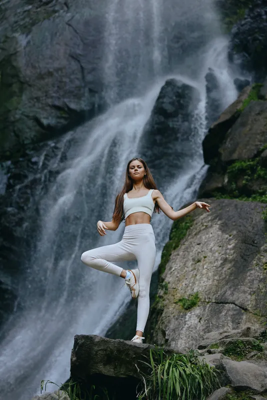 Young woman practicing yoga pose outdoors in front of a majestic waterfall, wearing white activewear, showing balance and calmness in nature.
