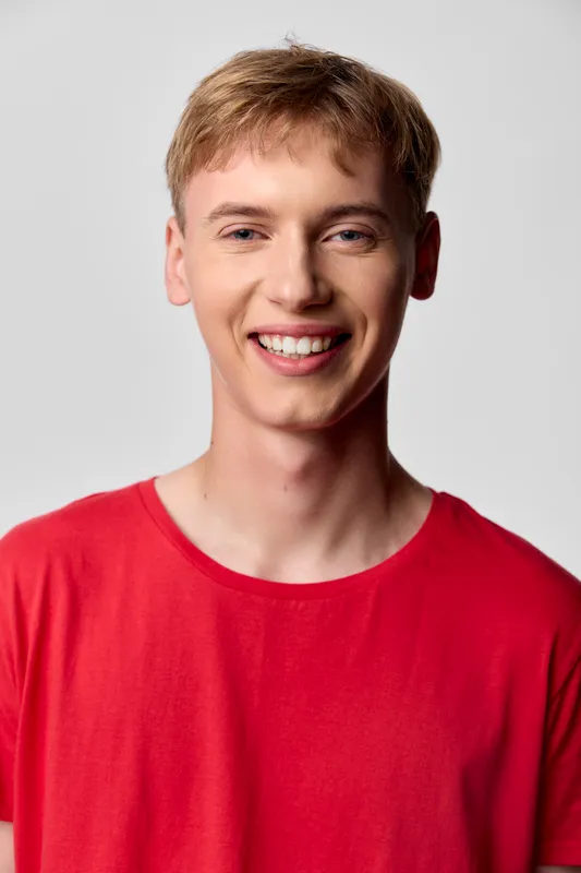 Smiling young man in a red shirt against a plain neutral backdrop, a clean casual headshot for lifestyle, fashion, or wellness campaigns, conveying approachable energy and friendly, confident