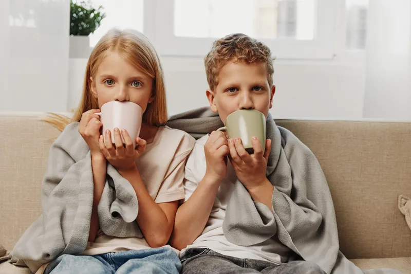 Two children wrapped in blanket drinking hot drinks on beige sofa at home. Cozy indoor moment with warm light and casual clothing for family comfort concept.