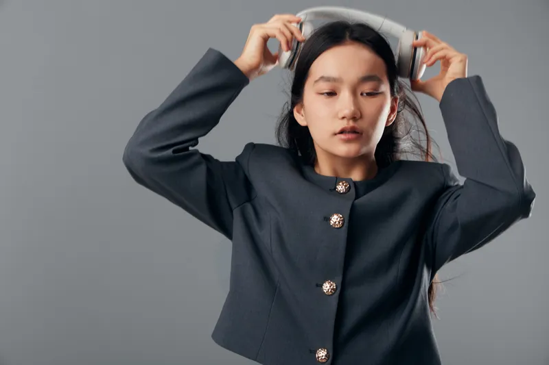 Woman with long dark hair wears stylish black jacket putting on white headphones isolated on gray background. Portrait of female listening to music with closed eyes and focused expression.