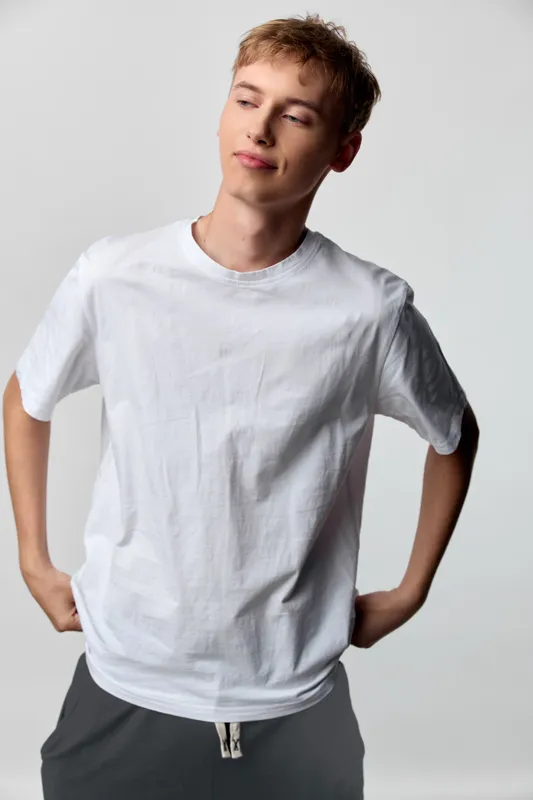 Young man wearing a white t shirt stands with hands on hips in a studio, relaxed pose, clean lighting, casual fashion style and a minimalist backdrop.