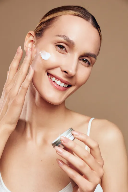 A smiling woman applies moisturizer cream on her cheek in a bright studio. Concept for skincare, beauty routine, healthy skin, and self care at home.