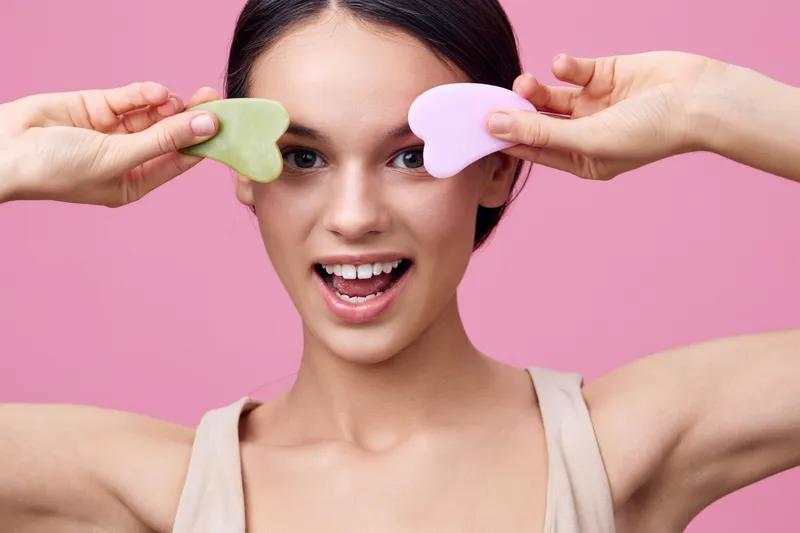 Young woman holding heart shaped sponges, smiling cheerfully on a pink background, representing beauty, skincare, and self care concepts