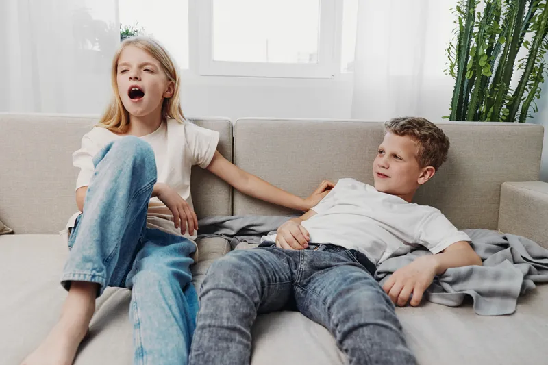 Two children relaxing on a sofa in a bright living room, one yawning and the other smiling while lying down. Casual home atmosphere with natural light and cozy setting.