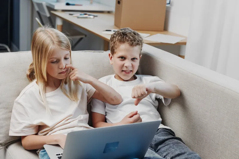 Two children sitting on a couch with a laptop, a boy showing a thumbs down, girl looking thoughtful. Concept of digital frustration and sibling interaction.