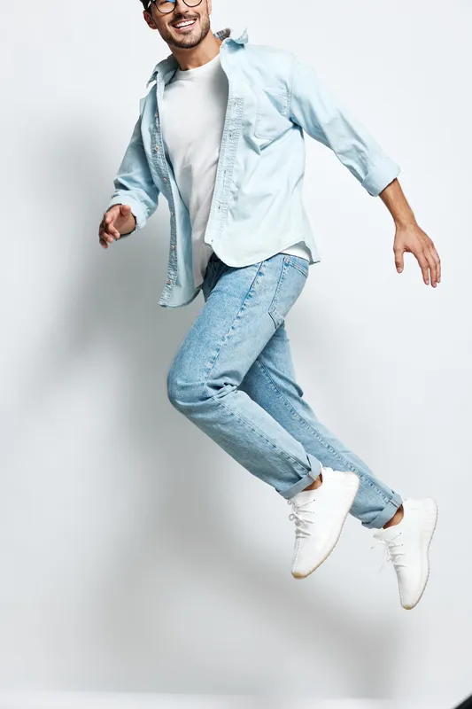 Full body joyful smiling young man in casual light blue outfit jumps in the air against a plain light background, expressing happiness and energy. Lifestyle concept.