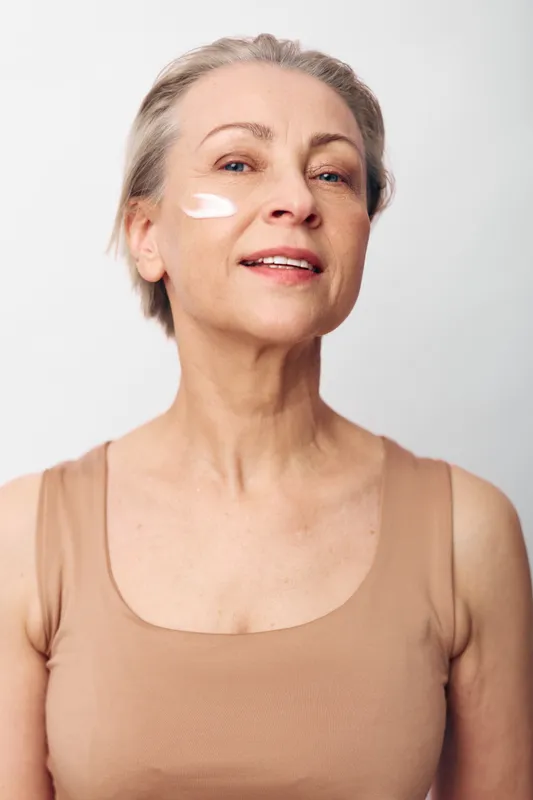 Mature woman with short grey hair applies white face cream on cheek, wearing beige tank top. Studio portrait on light background. Concept of skincare, aging beauty, and self-care routines.