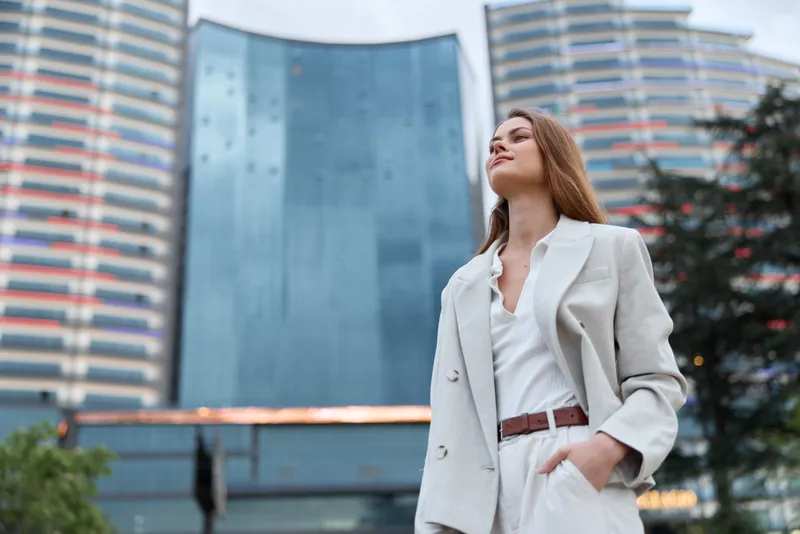 Confident woman in a business suit poses outdoors with modern architecture in the background, showcasing empowerment and professionalism in a contemporary setting.