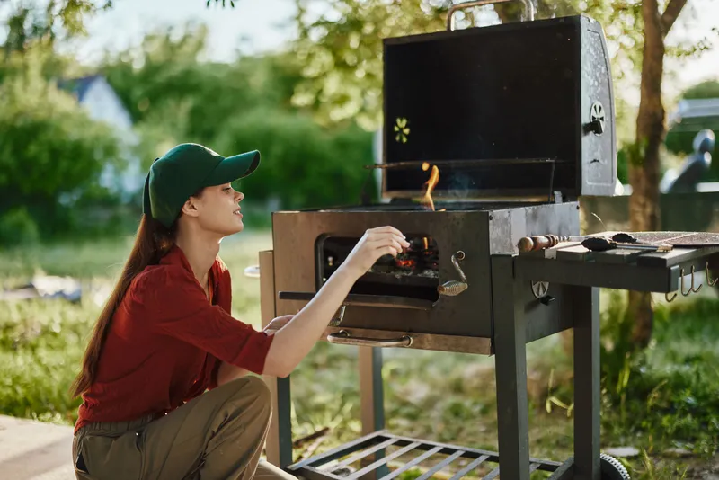 Young woman grilling outdoors in casual attire, enjoying the summer weather in a green garden setting with a barbecue grill and flames