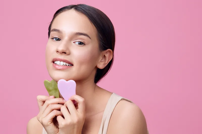 Young woman holding heart shaped skincare tools, smiling against a soft pink background, showcasing natural beauty and self care concept