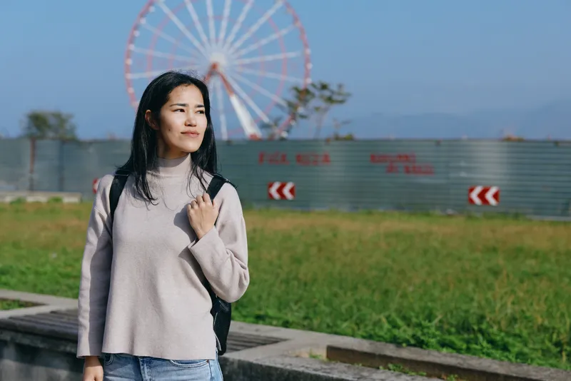 Woman with backpack stands along a path near a ferris wheel, outdoor park setting, casual clothing, relaxed mood, travel vibe, daytime light, emerging confidence.
