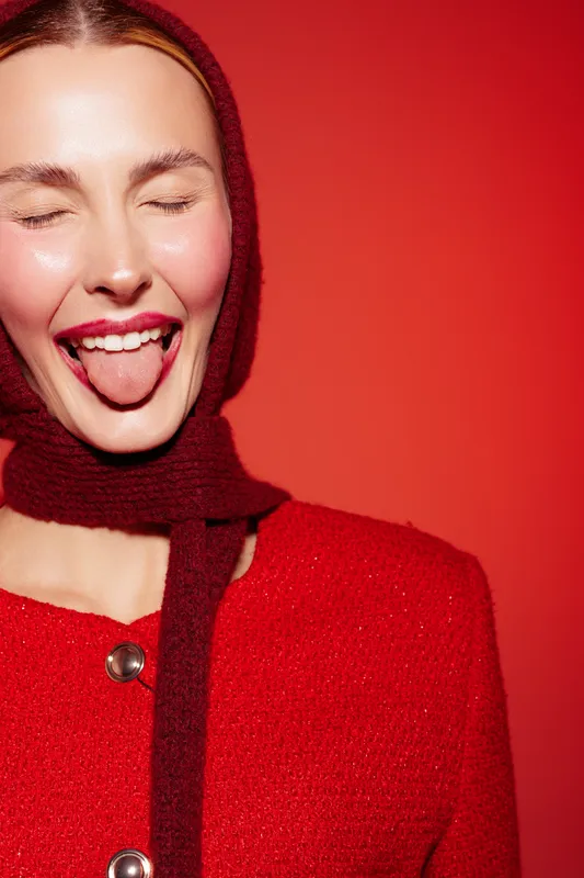 Young woman in red sweater and scarf sticks out tongue with closed eyes on red background. Playful expression and bright colors create fun and energetic mood.