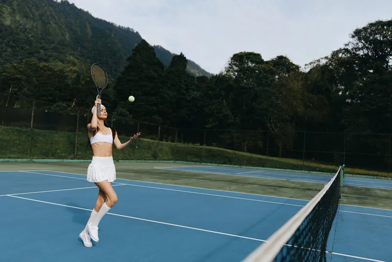 Young athletic woman in stylish tennis outfit practicing on sunny outdoor court, showcasing joy and determination in a scenic nature backdrop