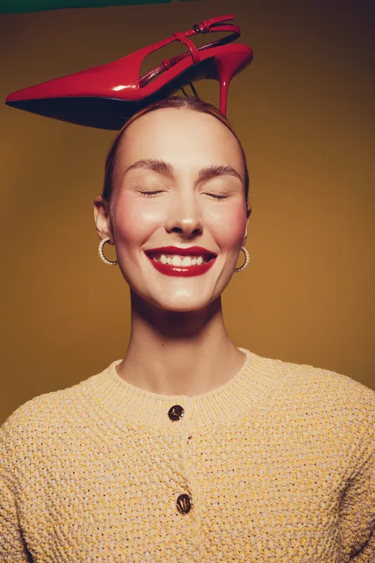 Smiling woman with closed eyes balances red high heel shoe on head wearing yellow knitted sweater and hoop earrings. Concept of fashion, joy, and playful mood in studio lighting.